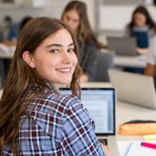 Portrait of smart university student in library with classmates in background. Close up face of school girl looking at camera while studying on computer. Smiling young woman looking behind while studying on laptop in university library.